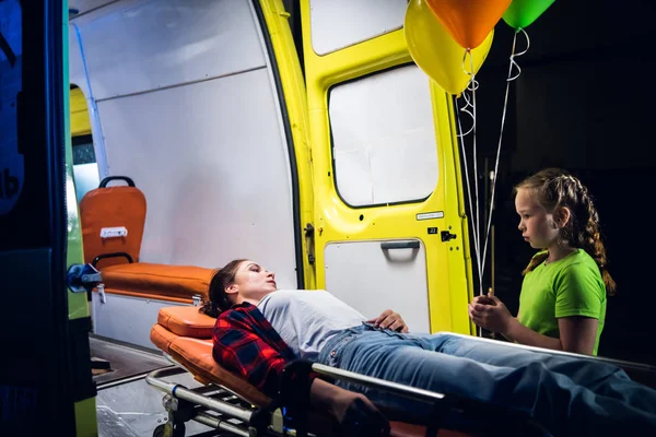 Girl stands beside a woman lying on stretcher, they look at each other in the ambulance car