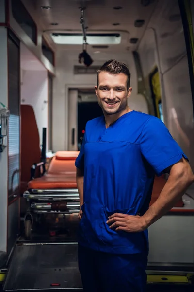 Ευτυχισμένος νοσοκόμος στέκεται και να δούμε κάμερα και χαμόγελο, ασθενοφόρο αυτοκίνητο στο παρασκήνιο — Φωτογραφία Αρχείου