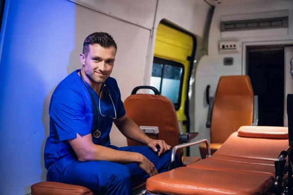 Un joven con uniforme médico se sienta en el coche de la ambulancia y sonríe —  Fotos de Stock
