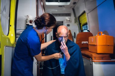 Young nurse in uniform puts her hand on man who sits in oxygen mask in blanket in the ambulance car