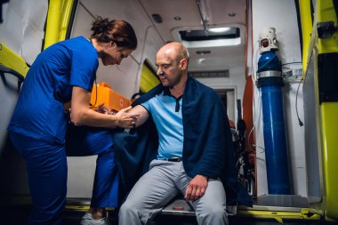 Man is sitting in blanket, nurse in blue uniform makes an injection in the ambulance car