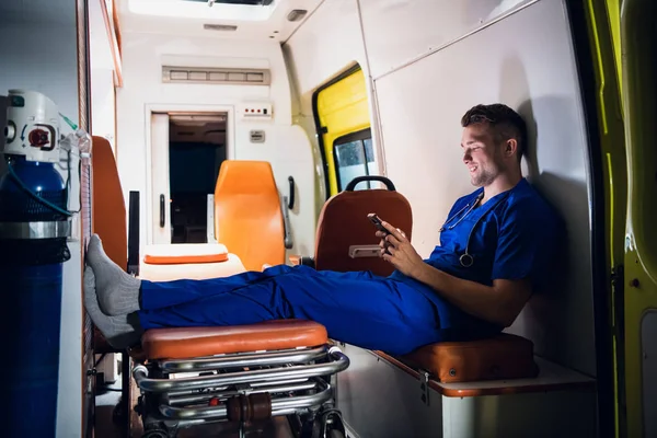 Молодой доктор сидит с телефоном в руках в машине скорой помощи ночью — стоковое фото