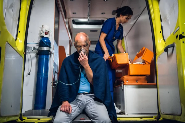 Hombre respirar a través de la máscara de oxígeno, enfermera joven en uniforme abre bolsa médica en el coche ambulancia — Foto de Stock
