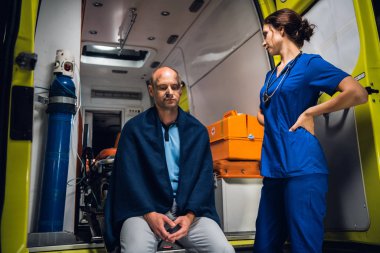 Üniformalı genç doktor ambulans arabasında battaniyeli bir adamla duruyor.