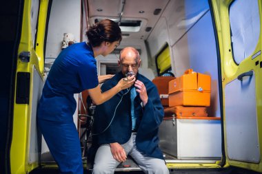 Young nurse in uniform puts oxygen mask on man sitting in blanket in the ambulance car