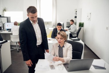 People work in the office, man in suit stands over the woman, who shows him the documents