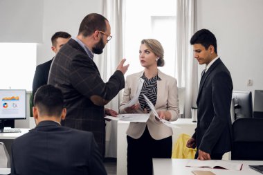 Chief in suit talk agressively with woman with documents in her hands, coworkers stand around them