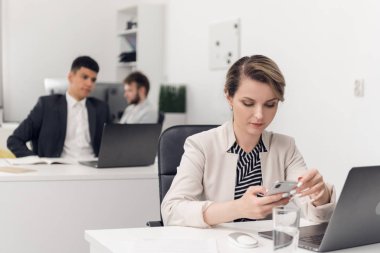 Young lady sits in the phone during working day, her colleages sit a a tables beside her