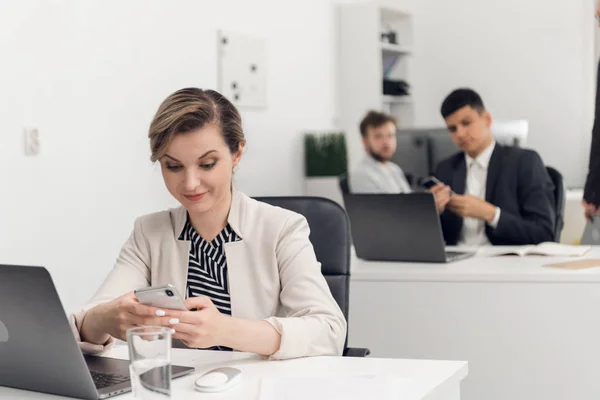 Młoda kobieta patrzy na telefon zaskakująco w biurze, koledzy siadają obok niej przy stoliku. — Zdjęcie stockowe