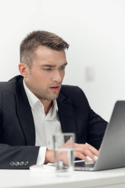 Focus on man in suit sitting at a table and typing on a laptop with serious face in the office