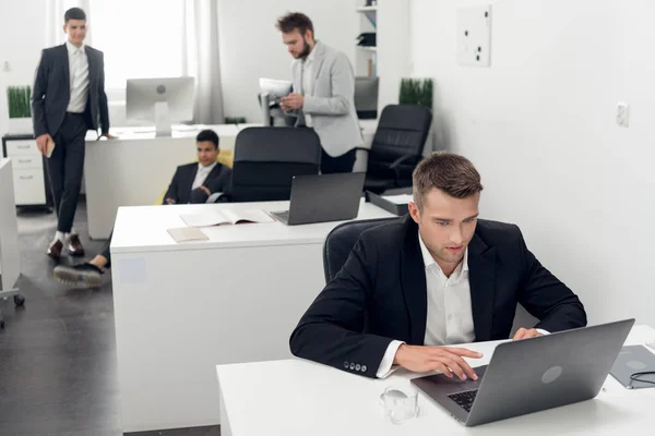 Человек сидит за столом и работает на ноутбуке в офисе, его коллеги говорят рядом с ним — стоковое фото