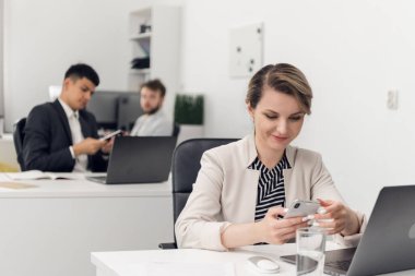 Ofiste çalışırken bir kadın telefonuna bakar ve gülümser. Meslektaşları yanında bir masa otururlar.