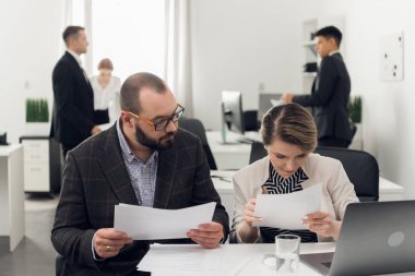 Takım elbiseli kadın ve erkek bir masada belgelerle oturuyor, diğer meslektaşlar onların yanında duruyor ve konuşuyorlar.