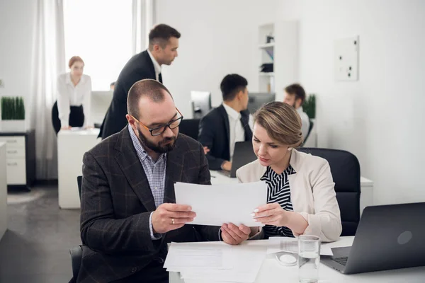 Женщина и мужчина сидят за столом с документами, рядом с ними работают другие коллеги — стоковое фото