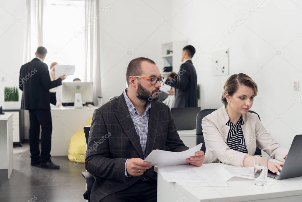 Focus on man with documents in his hands and woman sitting at a table and working on laptop, other workers are talking beside them in the office
