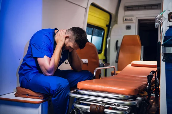 Üniformalı yorgun sıhhiyeci ambulansın içinde oturur ve ellerini başının üstüne koyar.