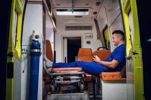 Paramedic in medical uniform sits with phone in his hands in the ambulance car at night — 图库照片