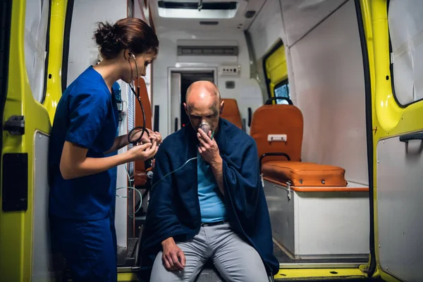 Enfermera de pie con estetoscopio y el hombre se sienta en la máscara de oxígeno en la manta en el coche ambulancia —  Fotos de Stock