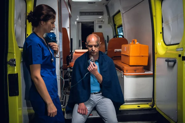 Молодая медсестра в форме стоит со стетоскопом, а мужчина сидит в кислородной маске в одеяле — стоковое фото