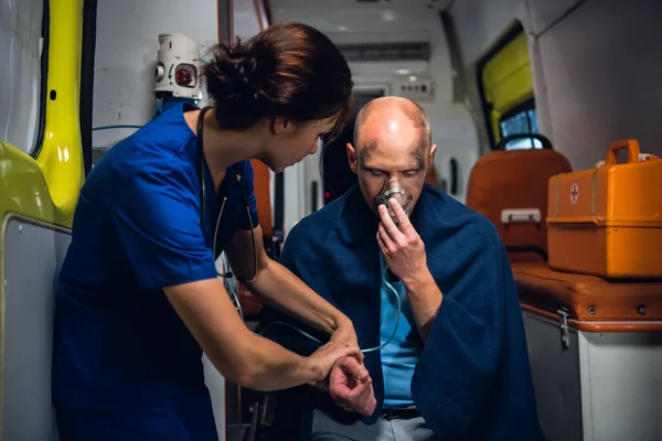 Молодая медсестра в форме держит руку человека, который сидит в кислородной маске в одеяле в машине скорой помощи — стоковое фото