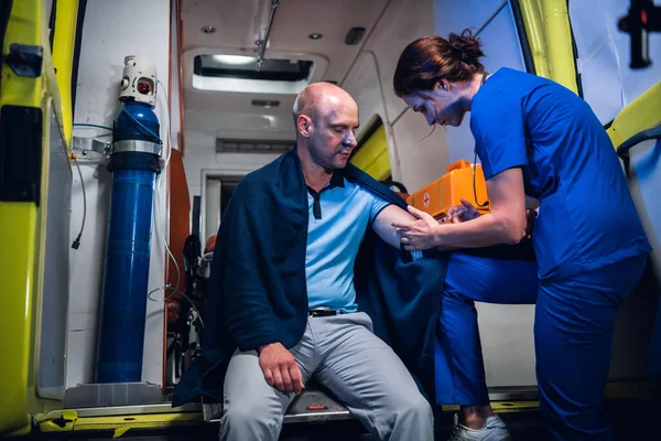 Мужчина сидит в одеяле, молодая медсестра в форме делает укол в машине скорой помощи — стоковое фото