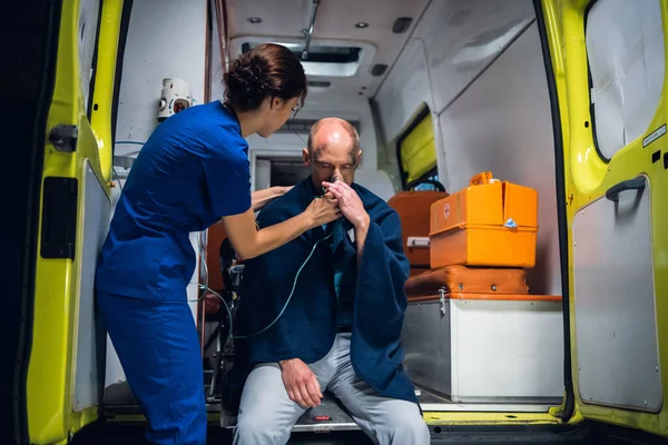 Enfermera joven en uniforme pone máscara de oxígeno en el hombre sentado en manta en el coche de la ambulancia —  Fotos de Stock
