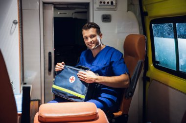 Tıbbi üniformalı sıhhiyeci ambulans vagonunda oturur ve tıbbi çantalarını toplar.