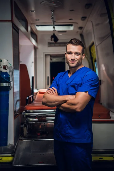 Un médico sonriente con uniforme médico se para sobre el coche de la ambulancia en el fondo —  Fotos de Stock