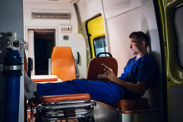 Парамедик в форме сидит с телефоном в руках в машине скорой помощи ночью — стоковое фото