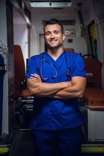 Doutor de uniforme fica e olha para a câmera sorrindo no carro da ambulância — Fotografia de Stock