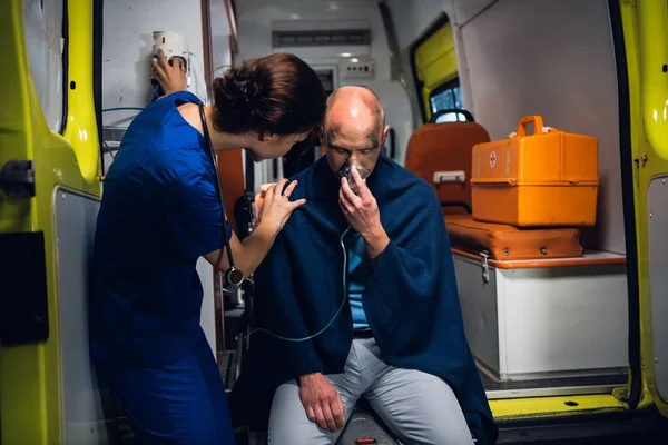 Wanita berseragam medis berdiri bersama pria yang duduk di dalam masker oksigen dengan selimut di mobil ambulans pada malam hari — Stok Foto