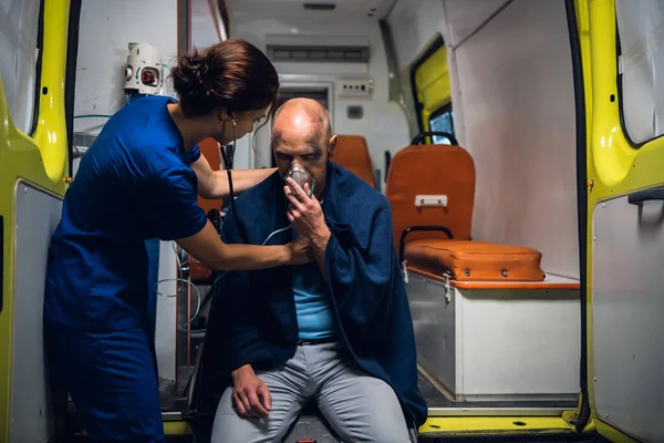 Enfermera con estetoscopio y hombre se sienta en máscara de oxígeno en manta en el coche ambulancia —  Fotos de Stock