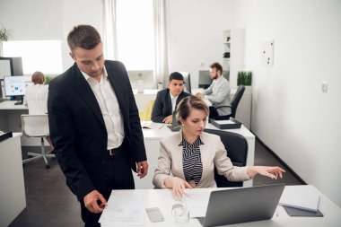 İnsanlar ofiste çalışıyor, takım elbiseli adam kadının başında duruyor ve belgelere bakıyor, kadın ellerini hareket ettiriyor.