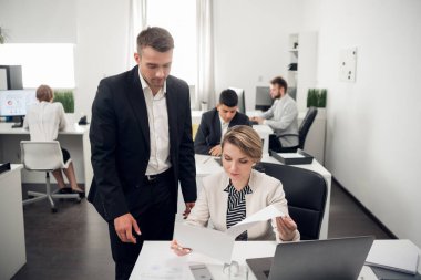 İnsanlar ofiste çalışıyor, genç yakışıklı adam ona belgeleri gösteren kadının yanında duruyor.