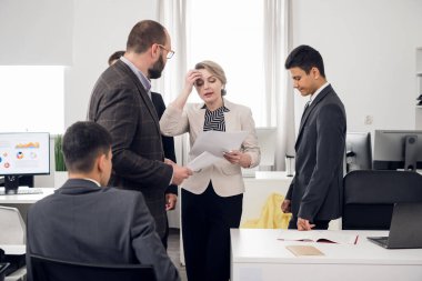 Elinde evrakları olan bir kadın şefine bir şeyi açıklar, iş arkadaşları yanlarında durur.