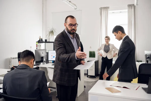 Serious chief talks to his young colleages and shows documents, coworkers stand around them in the office — 스톡 사진