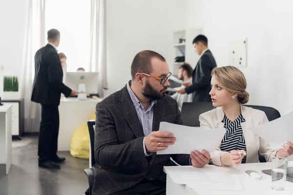 Focus on man in suit talking to a woman sitting at a table, other workers are talking beside them in the office