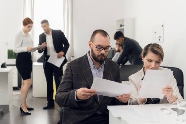Kadın ve erkek ellerinde belgelerle ofiste bir şeyler arıyorlar. Ofis çalışanları yanlarında konuşuyorlar.