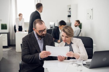 Kadın ve erkek bir masada belgelerle oturuyor ve onları tartışıyor, diğer meslektaşlar da onların yanında çalışıyor.