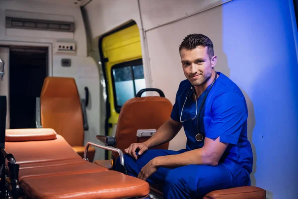 Jovem de uniforme senta-se no carro da ambulância e sorri — Fotografia de Stock