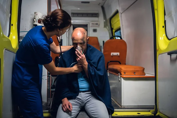Медсестра со стетоскопом и мужчина сидит в кислородной маске в одеяле в машине скорой помощи — стоковое фото