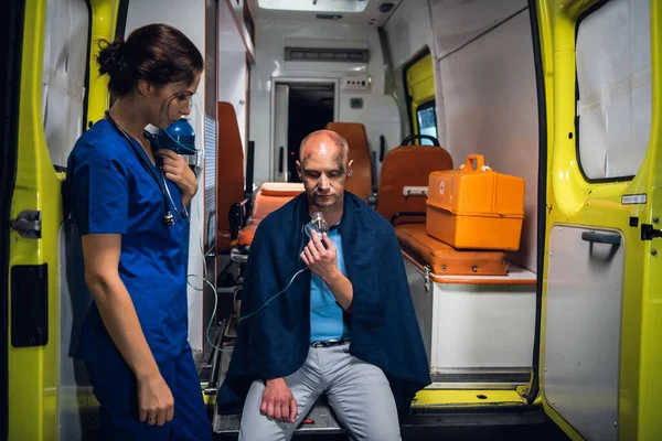 Медсестра в форме стоит со стетоскопом, а мужчина сидит в кислородной маске в одеяле в машине скорой помощи — стоковое фото