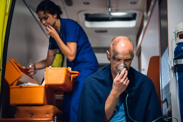 Hombre respirar a través de la máscara de oxígeno, enfermera joven abre algo tomado de la bolsa médica en el coche ambulancia —  Fotos de Stock