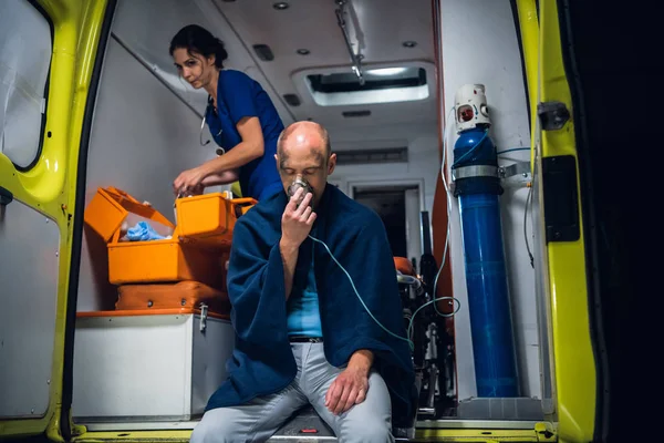 Mann in Decke atmet durch Sauerstoffmaske, junge Krankenschwester in Uniform packt Ausrüstung in medizinische Tasche — Stockfoto