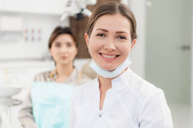 Kameraya gülümseyen kadın bir doktorun yakın plan fotoğrafı. Hastası arka planda kontrol edilmeyi bekliyor.