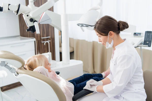 Um especialista odontológico pediátrico habilidoso está examinando os dentes de uma criança. A mulher está sentada perto da menina tentando estabelecer um contato com ela para fazê-la se sentir confortável — Fotografia de Stock