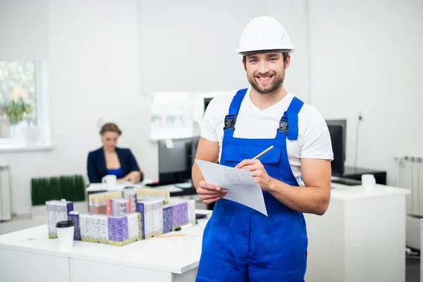 Een jonge bouwdirectie in een witte helm en blauwe overall maakt aantekeningen in het kantoor — Stockfoto