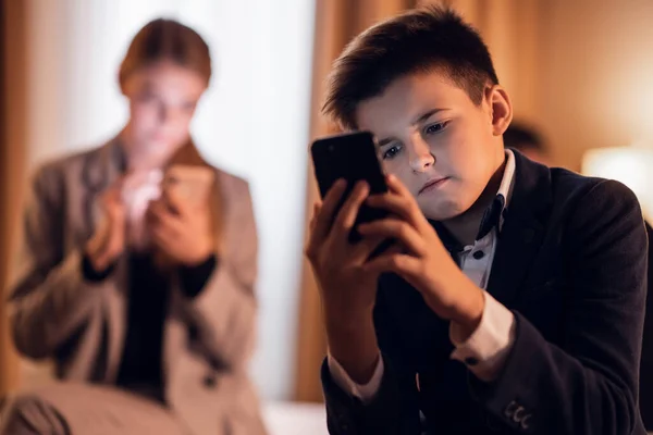 Un niño sentado y jugando en su teléfono inteligente, su hermana mayor en la parte posterior también mirando su teléfono — Foto de Stock