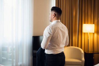 Beyaz gömlekli bir adam pencerenin önünde dikiz aynasında dikiz aynasından duruyor.