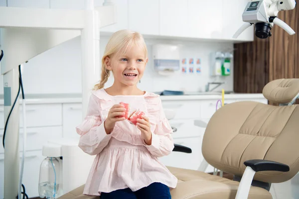 Un retrato de una pequeña paciente sentada en la silla del dentista con dentadura postiza en la mano y sonriendo con una sonrisa dentada — Foto de Stock